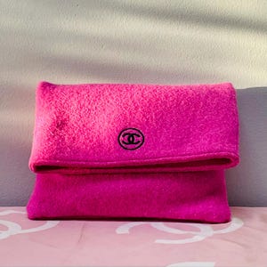 May include: A bright pink, folded clutch bag with a textured surface. The bag features a black logo on the front. The bag is made of a soft material and has a folded flap closure.