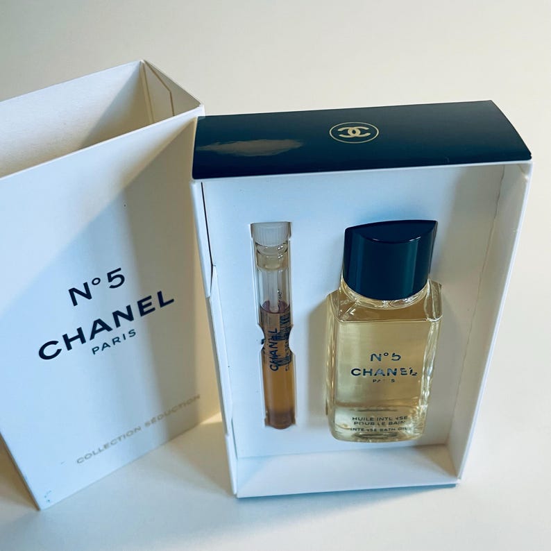 May include: A white box set containing a small perfume bottle and a sample vial of Chanel No. 5. The box has the Chanel logo and text. The perfume bottle has a black cap and a clear glass body with a yellow liquid.