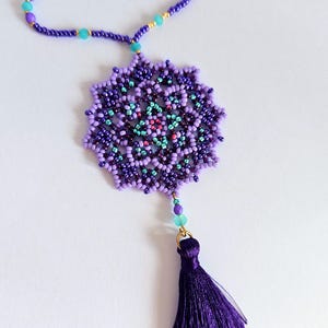 May include: A purple beaded necklace featuring a large, intricate mandala pendant and a purple tassel. The mandala is crafted with various shades of purple, blue, and green beads.