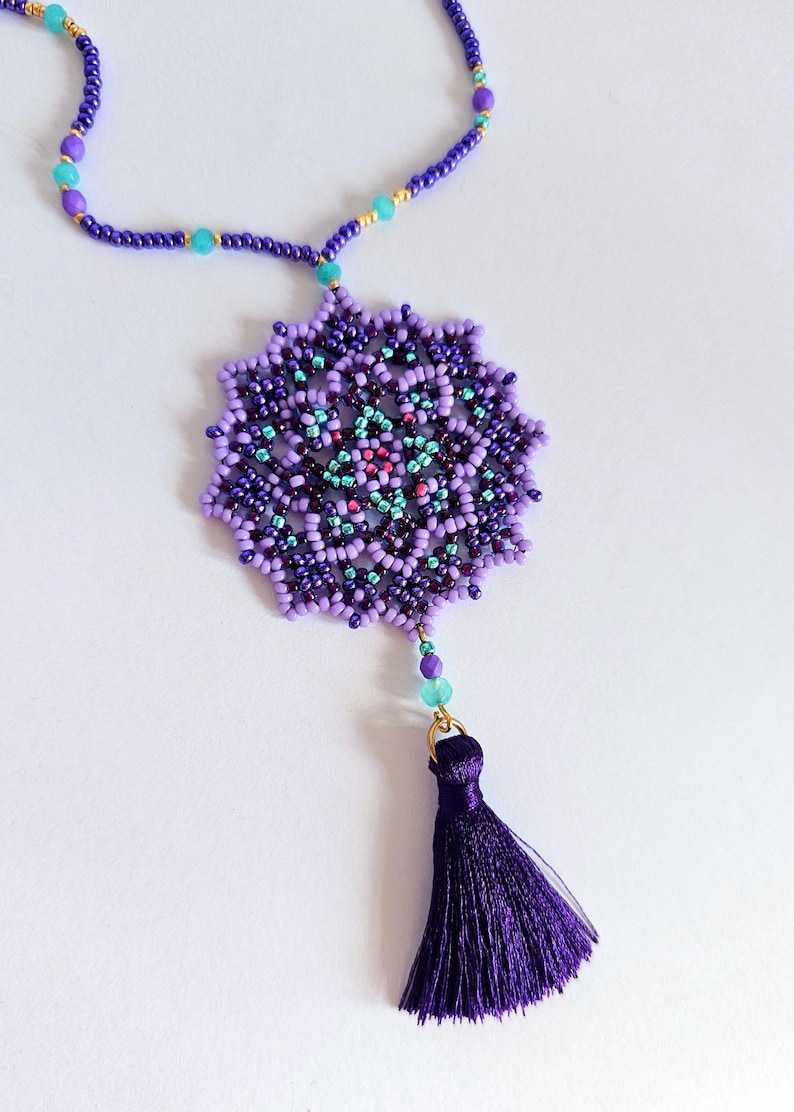May include: A purple beaded necklace featuring a large, intricate mandala pendant and a purple tassel. The mandala is crafted with various shades of purple, blue, and green beads.
