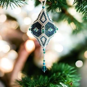 May include: A diamond-shaped beaded ornament featuring a geometric pattern in dark blue, teal, and metallic silver seed beads. The ornament is accented with small green crystals and terminates in a dangling strand of beads and a single teal crystal.