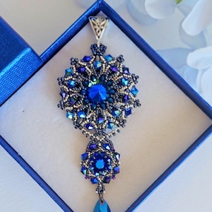 Puede incluir: Un colgante de cuentas azules y plateadas con un cristal en forma de lágrima. El colgante está en una caja de terciopelo azul.