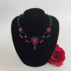 Può includere: Una collana di perline nere con accenti di cristallo rosso. La collana presenta un ciondolo a forma di cuore con un cristallo a forma di lacrima che pende.