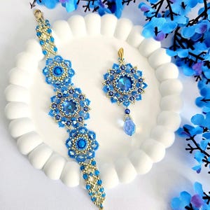 May include: A blue and gold beaded bracelet and pendant set. The bracelet and pendant feature floral designs with blue beads and gold accents. The pendant has a large blue central stone and a teardrop bead. The jewellery is displayed on a white plate, with blue floral decorations.