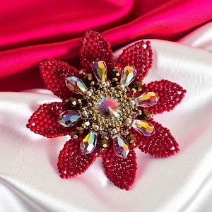 Può includere: Spilla a forma di fiore realizzata a mano con perline rosse, cristalli iridescenti e dettagli dorati. La spilla è posta su uno sfondo di tessuto di raso bianco e rosso, creando un contrasto sorprendente. Un accessorio floreale elegante.