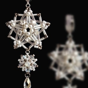 Può includere: Orecchino pendente argentato con un design a stella. L'orecchino presenta perline trasparenti, perle finte e cristalli. Una stella più piccola e un cristallo a forma di goccia pendono sotto. Lo sfondo è nero.