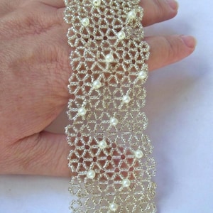 May include: A wide bracelet crafted from intricate, interlocking silver-toned seed beads arranged in a geometric, lace-like pattern. The design is accented with small, round, white faux pearls embedded at regular intervals throughout the structure.