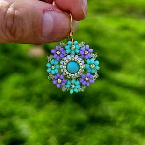 Può includere: Un paio di orecchini con un centro blu turchese circondato da un cerchio di perline bianche e un anello di fiori blu turchese e viola.