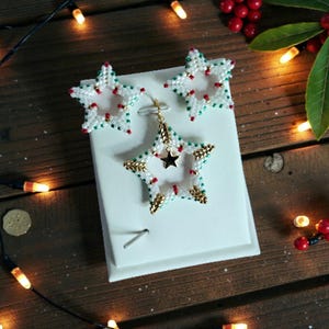 May include: A set of star-shaped earrings and a pendant. The earrings and pendant are white with red and green accents, and gold-colored detailing. The pendant features a small black star in the center. The jewelry is displayed on a white surface.