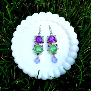 Puede incluir: Un par de pendientes colgantes con un dise&ntilde;o en capas. Los pendientes presentan piedras preciosas moradas y verdes, acentuadas con peque&ntilde;as cuentas en oro, turquesa y morado. Cada pendiente tiene un dije en forma de l&aacute;grima en la parte inferior y est&aacute; engarzado en un gancho plateado.