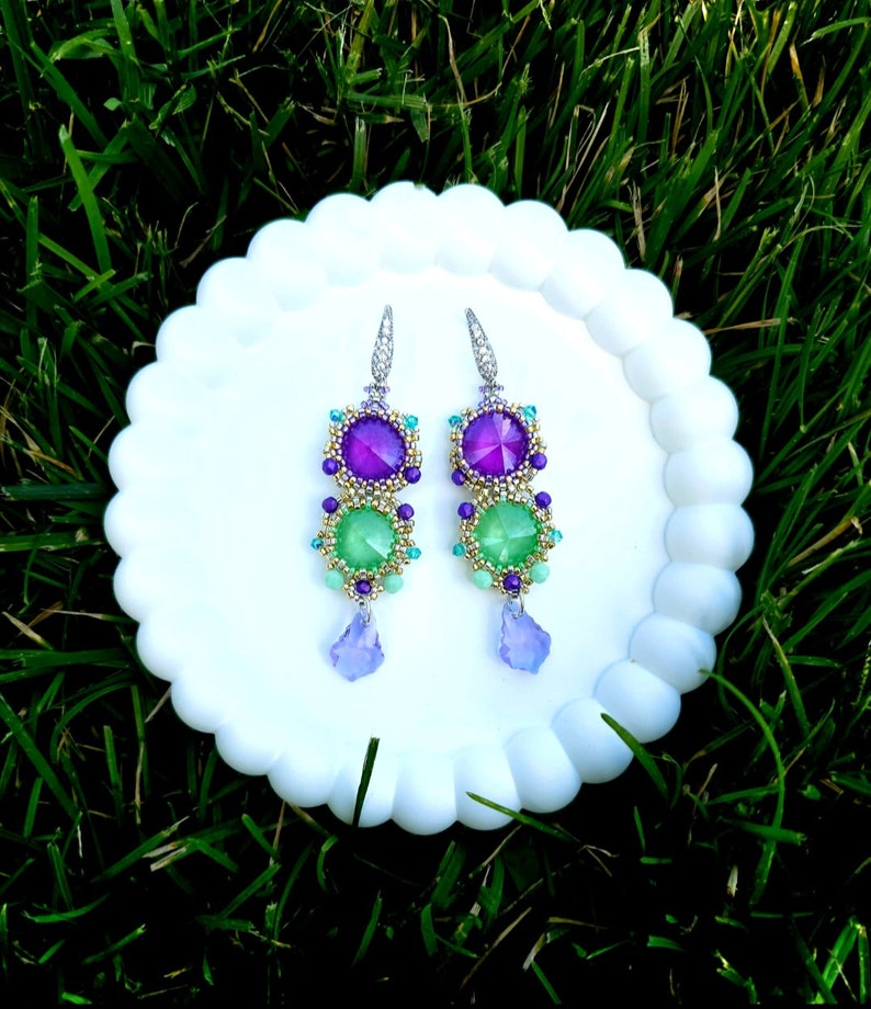 Puede incluir: Un par de pendientes colgantes con un dise&ntilde;o en capas. Los pendientes presentan piedras preciosas moradas y verdes, acentuadas con peque&ntilde;as cuentas en oro, turquesa y morado. Cada pendiente tiene un dije en forma de l&aacute;grima en la parte inferior y est&aacute; engarzado en un gancho plateado.