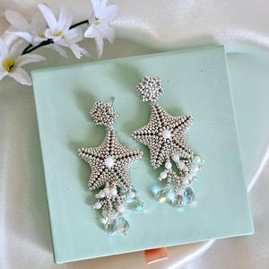 May include: A pair of silver starfish earrings with white pearl and crystal accents. The earrings are beaded and have a post backing. They are displayed on a light blue box with white flowers in the background.
