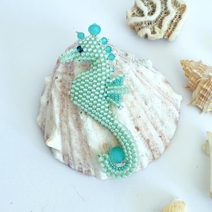Può includere: Un cavalluccio marino di perline turchese e acquamarina, con una perla turchese arrotondata sulla coda e sulla testa. Il cavalluccio marino è esposto su una grande conchiglia bianca, con altre conchiglie e coralli sullo sfondo.