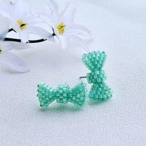 Puede incluir: Un par de pendientes de botón en forma de lazo de color verde menta. Los pendientes están hechos de pequeñas cuentas cuadradas y se muestran sobre una superficie blanca con flores blancas en el fondo. Los pendientes tienen un poste y un respaldo.
