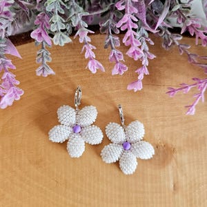 May include: A pair of white beaded flower earrings with a purple centre, set against a wooden background with purple flowers. The earrings have a silver hoop closure and are designed in a floral pattern.