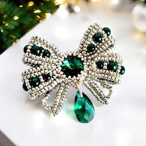 Può includere: Un broche a forma di fiocco in perline argentate e verdi con un grande cristallo verde al centro e un cristallo verde più piccolo a forma di lacrima che pende dalla parte inferiore.