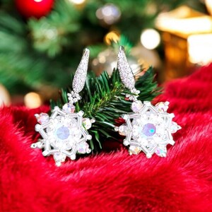 Op de afbeelding: Een paar zilveren sneeuwvlok oorbellen met heldere, iriserende kristallen die hangen aan een groene kerstboomtak. De oorbellen liggen op een rood, pluche oppervlak.