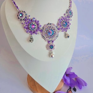 Op de afbeelding: Een paarse en zilveren kralenketting met een bloemmotief. De ketting heeft een grote, centrale hanger met een cluster van iriserende kristallen.