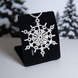 May include: A white beaded snowflake ornament displayed on a black velvet stand. The snowflake design features intricate detailing with small, round beads. The background includes a blurred winter scene with snow-covered trees.