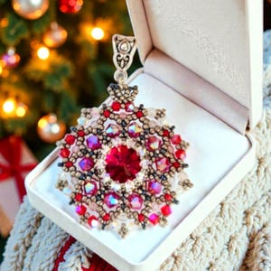 Op de afbeelding: Een met kralen versierde kerstversiering in een witte geschenkdoos. De versiering heeft een centrale rode kristal omringd door rode, roze en heldere kralen. De versiering zit in een witte geschenkdoos, met een kerstboom en lichten op de achtergrond.