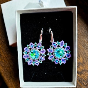 May include: A pair of silver dangle earrings with a teal and purple beaded flower design. The earrings are in a white box with black velvet lining.