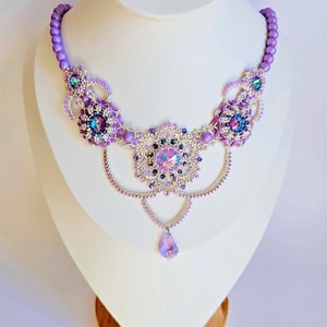 Puede incluir: Un delicado collar de cuentas moradas y plateadas con un gran colgante de cristal iridiscente. El collar presenta un diseño floral con múltiples capas de cuentas.