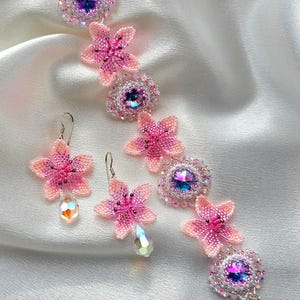 May include: A pink and white beaded bracelet and earrings set with crystal accents. The bracelet features a series of beaded flowers with a crystal centre. The earrings are dangle style with a beaded flower and a crystal drop.