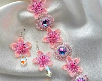 May include: A pink and white beaded bracelet and earrings set with crystal accents. The bracelet features a series of beaded flowers with a crystal center. The earrings are dangle style with a beaded flower and a crystal drop.
