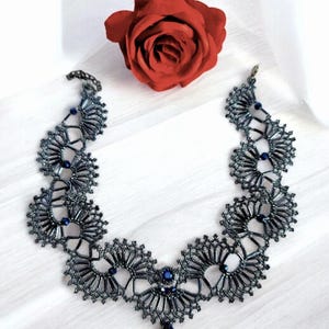 May include: A dark blue beaded necklace with a floral design and a teardrop pendant. The necklace is displayed on a white surface next to a red rose. The necklace is a unique piece of jewelry.