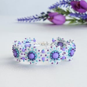 May include: A beaded bracelet featuring a floral design with white, light blue, and purple beads. The bracelet has a silver clasp and is set against a soft background with lavender flowers.