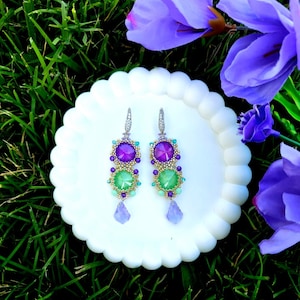 Puede incluir: Un par de pendientes colgantes con piedras preciosas moradas y verdes, presentados sobre un plato blanco con bordes festoneados. Los pendientes presentan una combinaci&oacute;n de piedras redondas y en forma de l&aacute;grima, con detalles dorados.