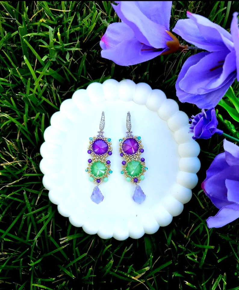 Puede incluir: Un par de pendientes colgantes con piedras preciosas moradas y verdes, presentados sobre un plato blanco con bordes festoneados. Los pendientes presentan una combinaci&oacute;n de piedras redondas y en forma de l&aacute;grima, con detalles dorados.