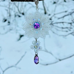 Peut inclure: Pendentif artisanal en perles blanches en forme de flocon de neige, orné d'un cristal violet central et d'une goutte de cristal violet en forme de goutte. Le pendentif est suspendu à une chaîne argentée.