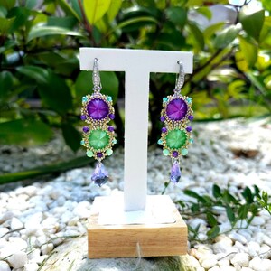 Puede incluir: Un par de pendientes colgantes con un dise&ntilde;o vibrante. Cada pendiente presenta una gran gema p&uacute;rpura en la parte superior, seguida de una gema verde y un cristal p&uacute;rpura colgante. Los pendientes est&aacute;n engarzados en metal plateado con peque&ntilde;as cuentas de colores.