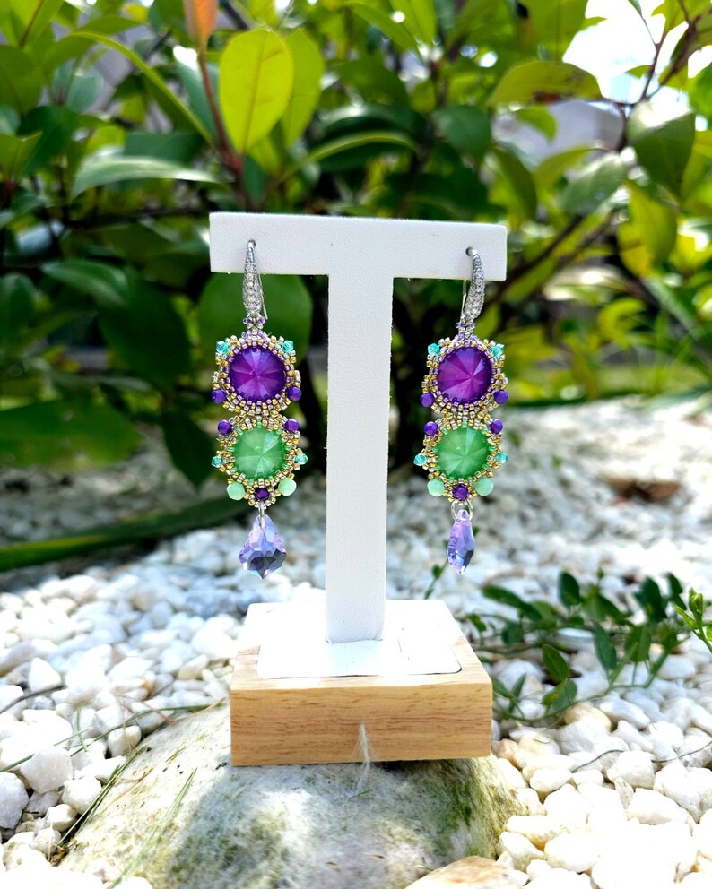 Puede incluir: Un par de pendientes colgantes con un dise&ntilde;o vibrante. Cada pendiente presenta una gran gema p&uacute;rpura en la parte superior, seguida de una gema verde y un cristal p&uacute;rpura colgante. Los pendientes est&aacute;n engarzados en metal plateado con peque&ntilde;as cuentas de colores.
