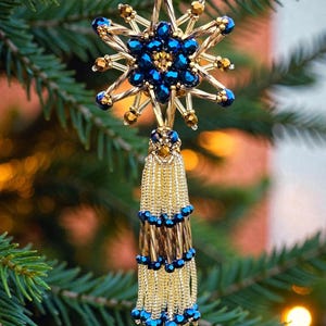 May include: A handcrafted Christmas tree ornament. The ornament features a starburst design with gold-coloured metal and blue and gold beads. A gold tassel hangs below the starburst, with blue and gold beads. The ornament is hanging on a green Christmas tree.