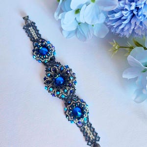 May include: A handcrafted bracelet featuring a dark blue beaded design with three large, round, faceted blue gemstones. The bracelet is adorned with smaller blue and silver beads, and is set against a white background with blue flowers.
