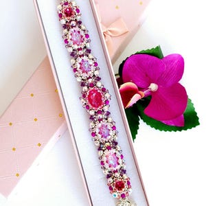 以下が含まれることがあります： ピンクとパープルのビーズブレスレットにシルバーの留め具が付いており、白い背景のピンクのギフトボックスに入っています。