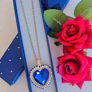 Może przedstawiać: Srebrny naszyjnik z dużym, niebieskim wisiorkiem w kształcie serca, otoczonym przezroczystymi kryształami. Wisiorek jest prezentowany w niebieskim pudełku, z pasującym małym pudełkiem i czerwonymi różami w tle. Łańcuszek jest delikatny.