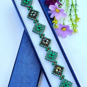 Puede incluir: Una pulsera de cuentas con una serie de motivos en forma de diamante interconectados en tonos turquesa, verde y bronce. La pulsera alterna entre cuentas verdes sólidas y cristales facetados iridiscentes, todo dentro de una caja azul.