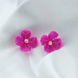 Könnte beinhalten: Ein Paar magentafarbene, mit Perlen besetzte Blumenohrringe mit einem Perlenmittelpunkt. Die Ohrringe sind vor einem weißen, strukturierten Hintergrund platziert. Das florale Design besteht aus kleinen, eng aneinander liegenden Perlen.