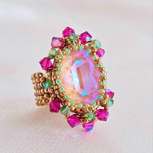Könnte beinhalten: Handgefertigter Ring mit einem großen, birnenförmigen, irisierenden rosa Edelstein. Der Edelstein ist von einem goldfarbenen Perlenrahmen umgeben, der mit kleineren rosa und grünen Kristallen akzentuiert ist. Das Ringband ist ebenfalls goldfarben und mit Perlen besetzt.