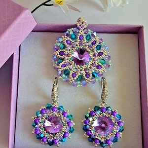 Pode incluir: Um conjunto de brincos e um colar com pingente de contas roxas, turquesa e prateadas. As joias apresentam um grande cristal transparente no centro de cada peça.