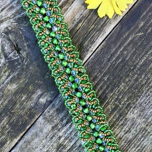Puede incluir: Un brazalete de cuentas verdes y doradas, con un patrón repetitivo. El brazalete mide aproximadamente 18 cm de largo. Una flor amarilla está en el fondo, sobre una superficie de madera envejecida.