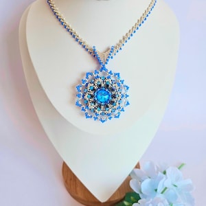 Op de afbeelding: Een blauwe en zilveren kralenketting met een grote blauwe kristallen hanger. De hanger is omgeven door kleinere blauwe en zilveren kralen.