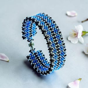 May include: A beaded bracelet with a blue, black, and silver pattern. The bracelet is made of small beads woven together to create a textured surface. The bracelet is approximately 1 inch wide and features a clasp closure.