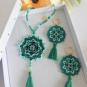 May include: A set of green and blue beaded jewellery with tassel accents. The necklace and earrings feature a mandala design with red accents. The jewellery is displayed in a white box.