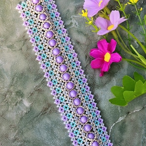 Könnte beinhalten: Ein Armband aus Perlen mit einer zentralen Reihe aus lavendelfarbenen Perlen, umgeben von weißen, hellblauen und lavendelfarbenen Perlen. Das Armband ist etwa 18 cm lang. Lila und gelbe Blumen sind ebenfalls zu sehen.
