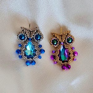 以下が含まれることがあります： 2つのビーズで作られたフクロウのイヤリング。1つは青いアクセントと大きな涙型の青いクリスタルが付いたシルバーです。もう1つは紫色のアクセントと大きな涙型の虹色のクリスタルが付いたゴールドです。どちらもビーズのデザインです。