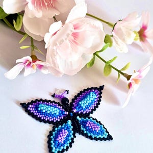 Peut inclure: Un papillon perlé avec des accents bleus, violets et noirs. Le papillon est composé de petites perles et présente un motif détaillé. Des fleurs roses et blanches sont en arrière-plan.